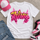 White T-shirt mockup with Kelsey in pink-and-yellow airbrush bubble script, styled beside jeans, pink sneakers, and a pink trucker hat.